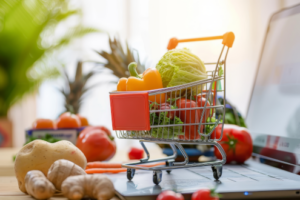 Symbolbild Online-Shopping: Kleiner Einkaufswagen mit Lebensmitteln steht auf Laptop